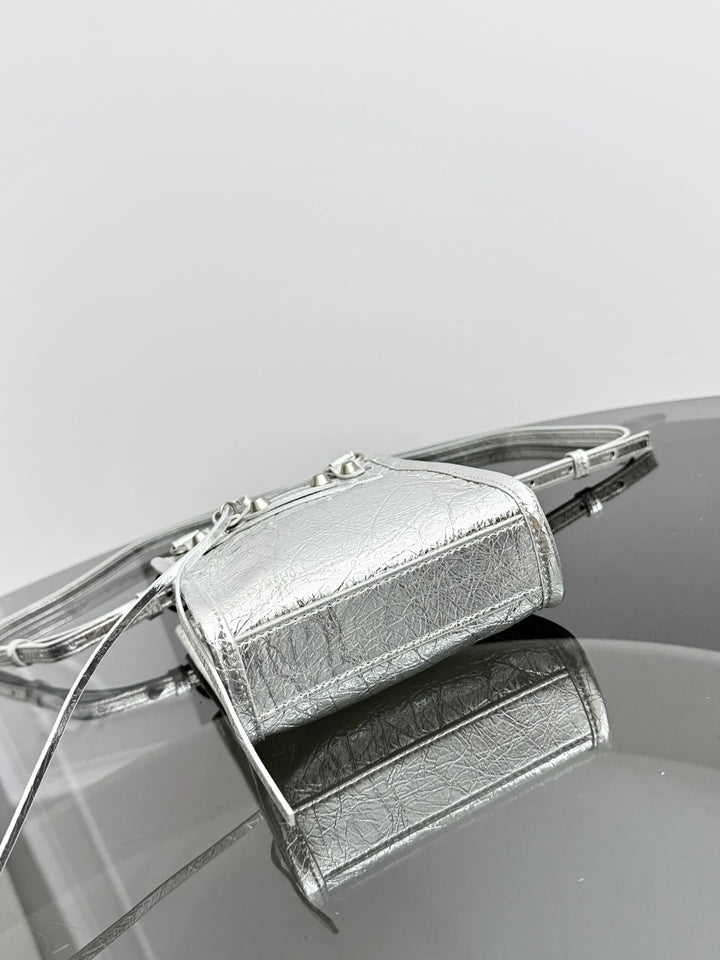 A metallic silver handbag with a textured leather finish, featuring a structured rectangular body, silver-tone metal frame details, and a thin strap, resting on a reflective dark surface against a light gray background.