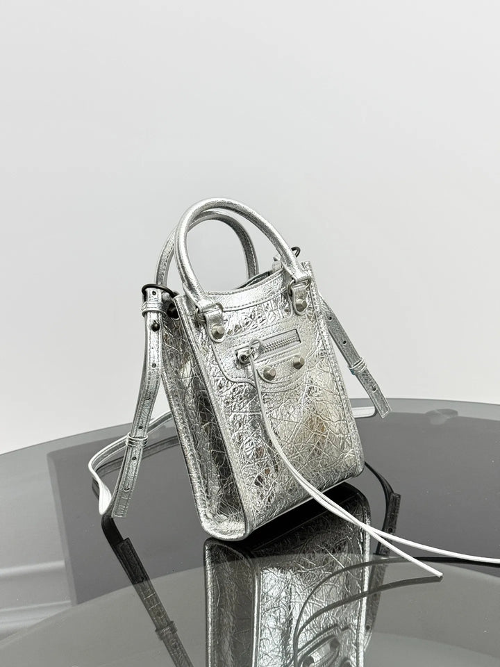 A metallic silver textured leather handbag with double top handles and a long shoulder strap, displayed on a reflective dark surface against a light gray background.