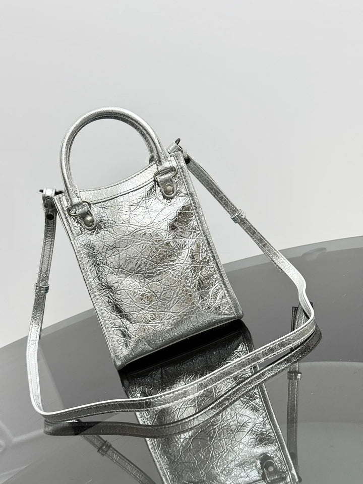 A metallic silver handbag with a textured leather finish, featuring a top handle, a detachable shoulder strap, and silver hardware, displayed on a reflective dark surface against a light gray background.