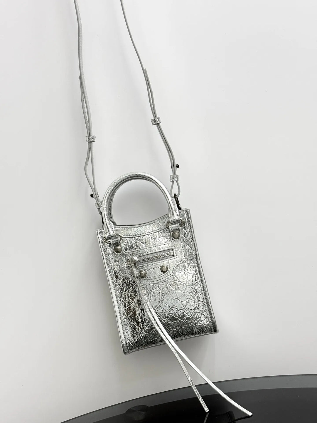 A metallic silver handbag with a textured crocodile pattern, featuring a top handle, a zippered front pocket, and a long thin shoulder strap, shown against a plain light gray background.