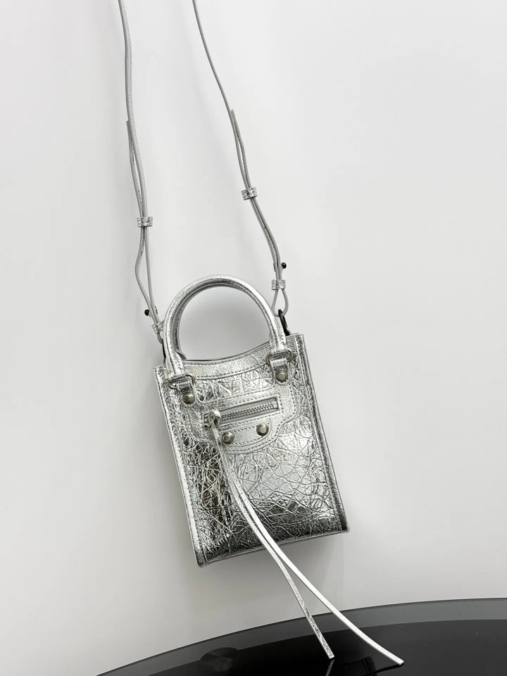 A metallic silver handbag with a textured crocodile pattern, featuring a top handle, a zippered front pocket, and a long thin shoulder strap, shown against a plain light gray background.