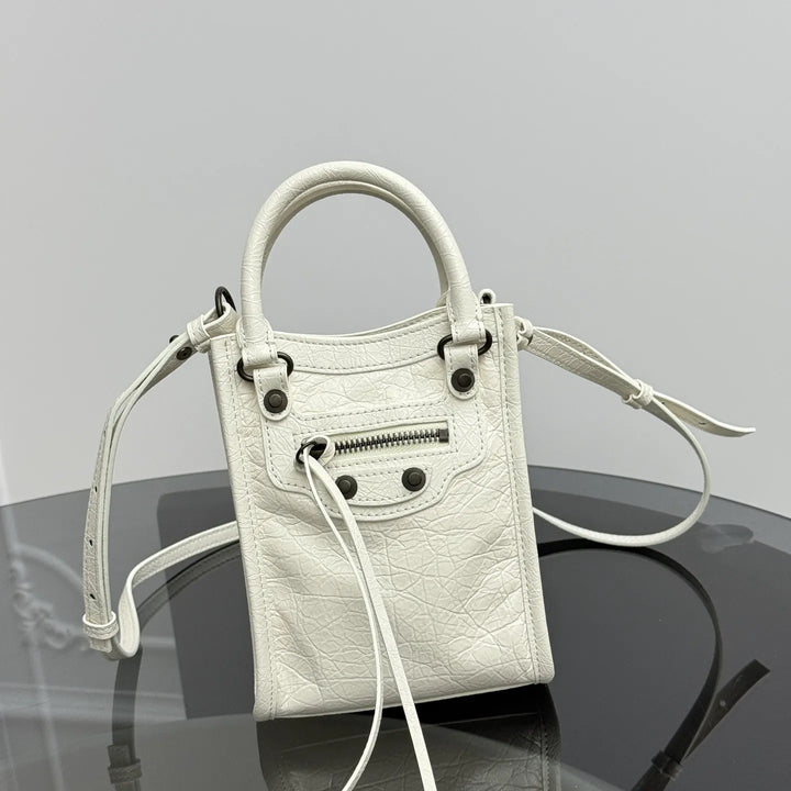 A white textured leather handbag with a top handle, front zip pocket, and shoulder strap, displayed on a reflective dark surface against a light gray background.