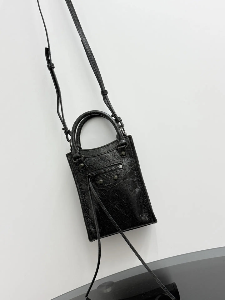 A black textured leather handbag with a top handle, a long shoulder strap, and a front flap detail, photographed against a plain white wall.