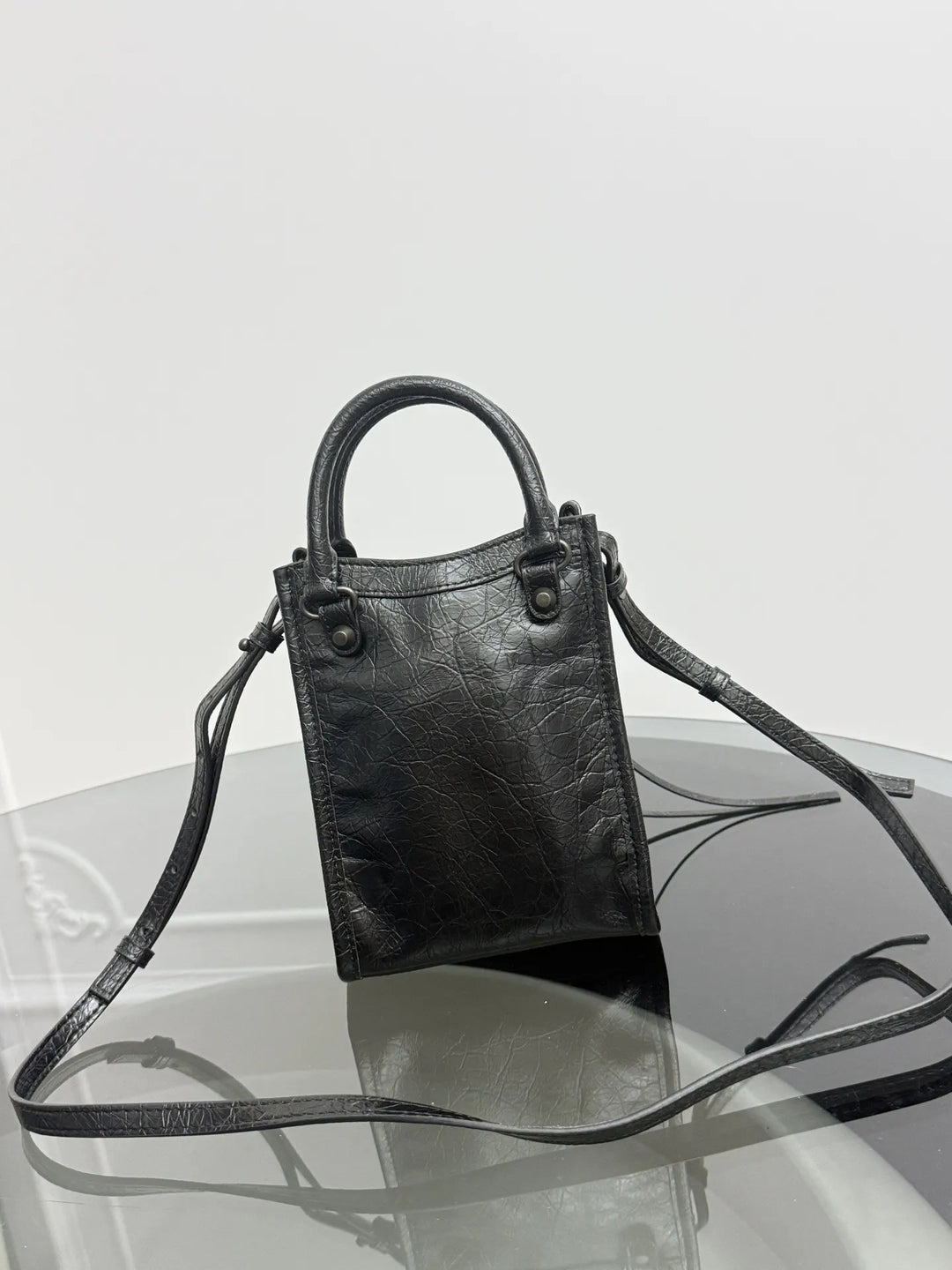 A small black leather handbag with a top handle and a long shoulder strap, displayed on a reflective glass surface against a light gray background.