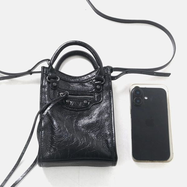 A small black leather handbag with a top handle and shoulder strap, shown on a white background next to a smartphone for scale.