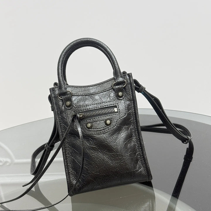 A black textured leather top-handle handbag with a detachable shoulder strap, displayed on a reflective glass surface against a light gray background.