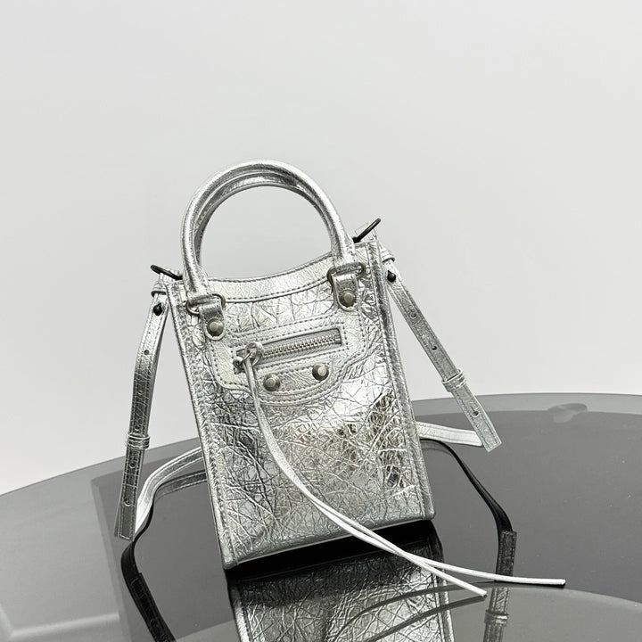 A metallic silver textured leather handbag with a top handle, front zip pocket, and long strap, displayed on a reflective dark surface against a light gray background.