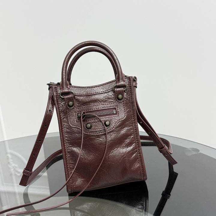A small structured leather handbag in dark reddish-brown with double top handles, a long shoulder strap, and metal hardware, photographed on a reflective dark surface against a light gray background.