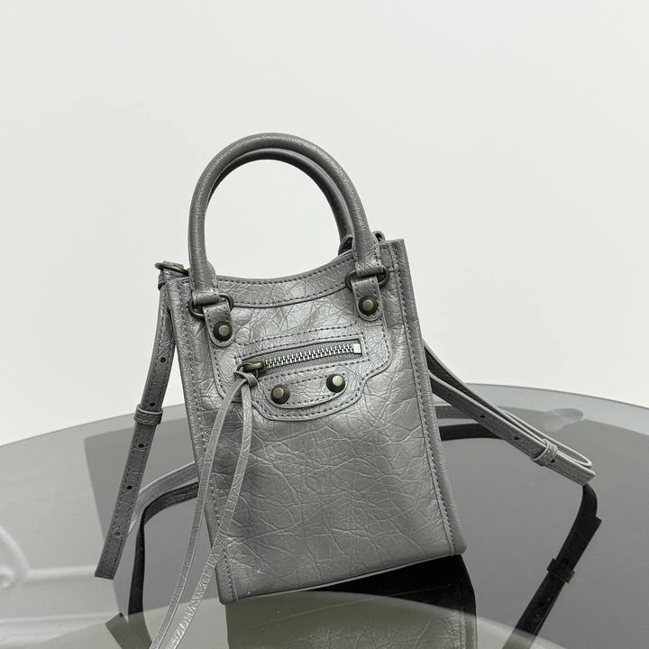 A small metallic silver leather handbag with a top handle, front zip pocket, and shoulder strap, displayed on a reflective dark surface against a light gray background.