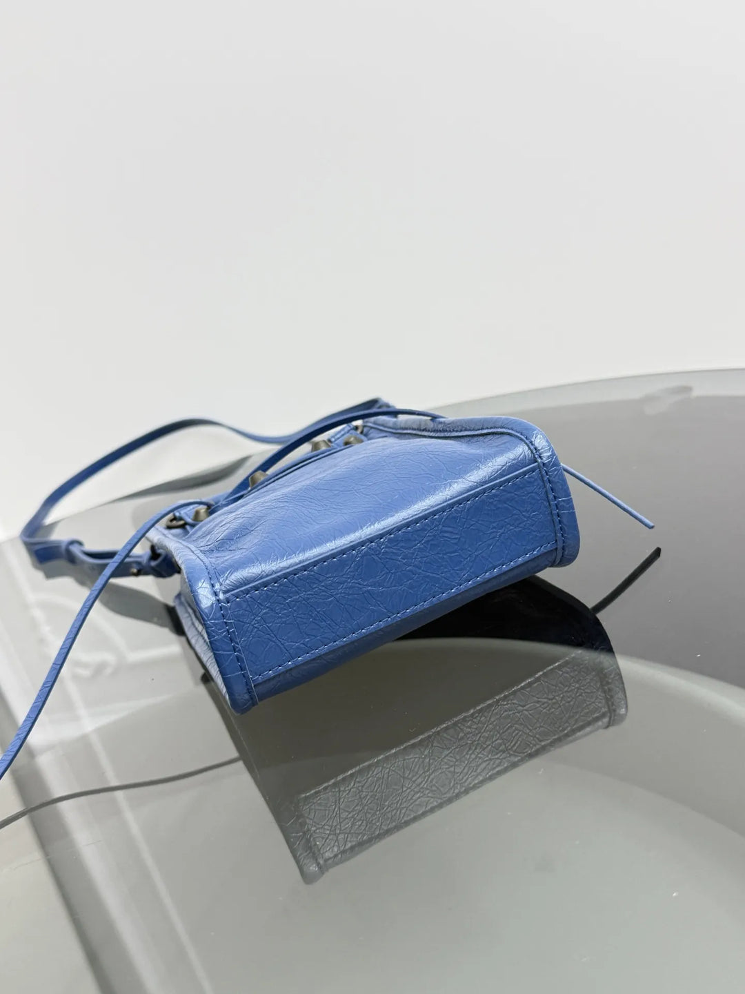 A small blue leather shoulder bag with a top zipper and thin strap, resting on a glossy gray surface against a light gray background.