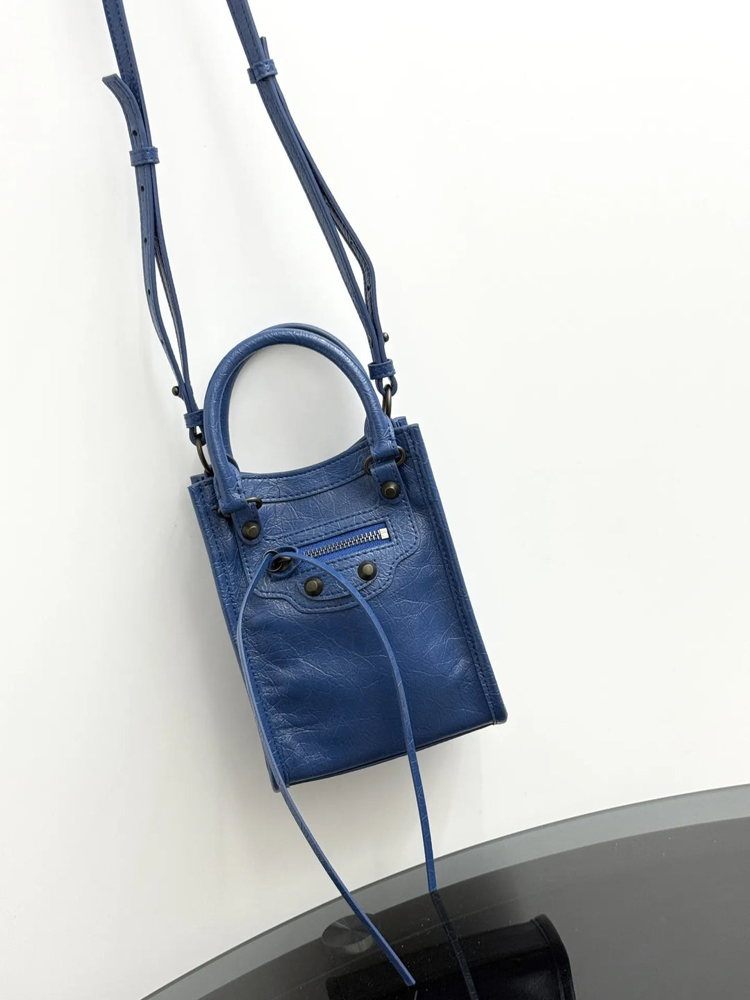 A blue leather top-handle handbag with a shoulder strap, displayed against a plain white wall.
