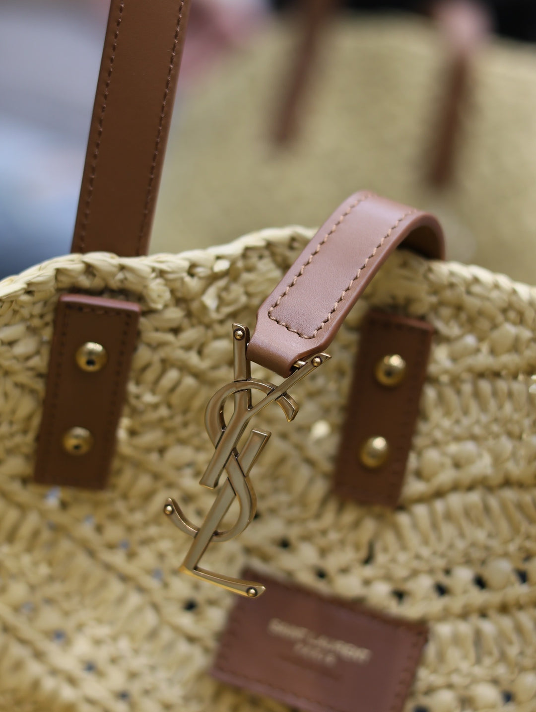 Close-up of a straw bag with a YSL logo charm, featuring brown leather straps and gold hardware.