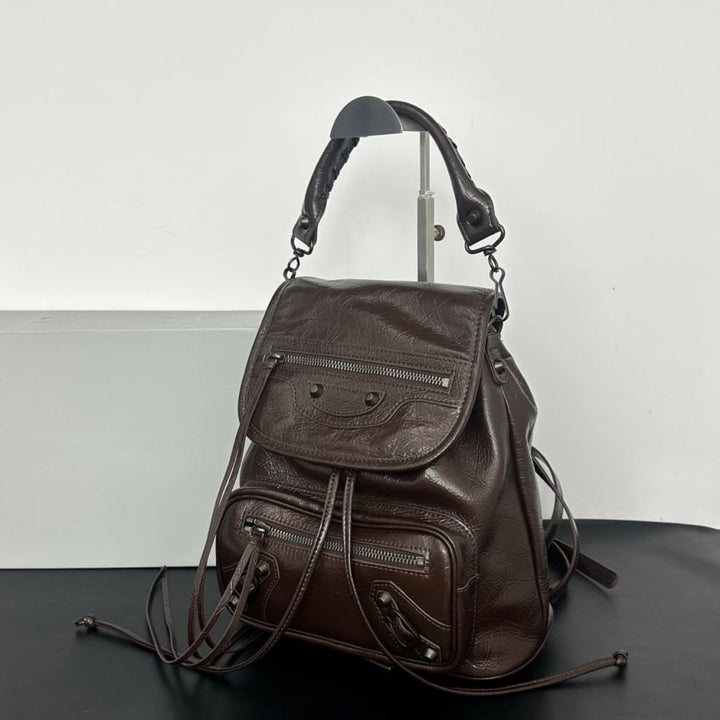 A dark brown pebbled leather backpack with multiple zippered pockets and a top handle, displayed on a stand against a white and gray background.