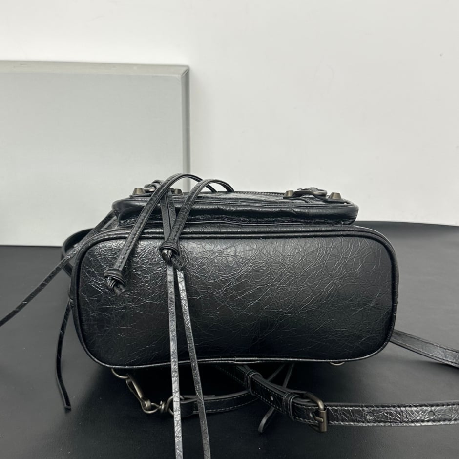 A black textured leather shoulder bag with a top zipper, hanging drawstring ties, and a matching shoulder strap resting on a dark surface against a plain light gray background.