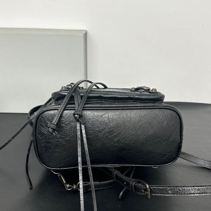 A black textured leather shoulder bag with a top zipper, hanging drawstring ties, and a matching shoulder strap resting on a dark surface against a plain light gray background.